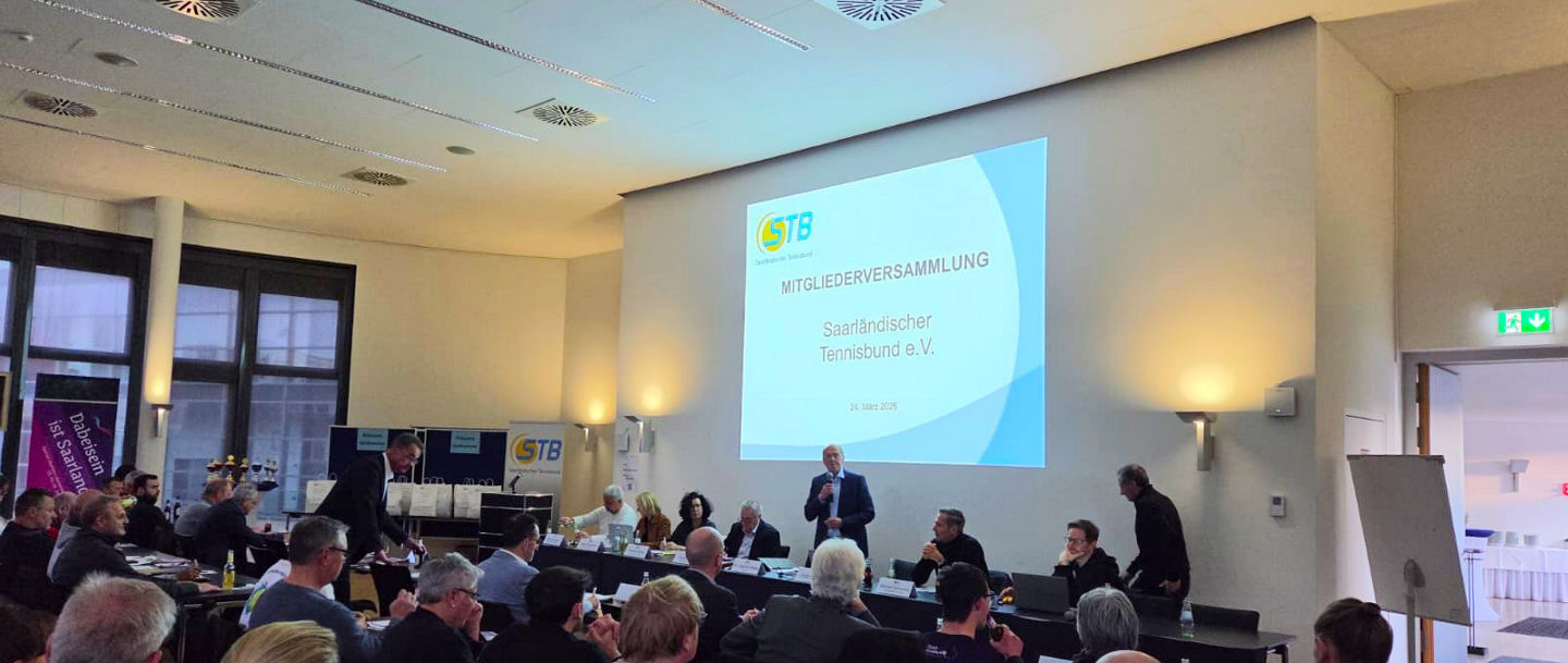 Blick in den Raum der Mitgliederversammlung, auf dem die Teilnehmenden von hinten und das Präsidium am anderen Ende des Raumes vor der Präsentation von vorne zu sehen sind