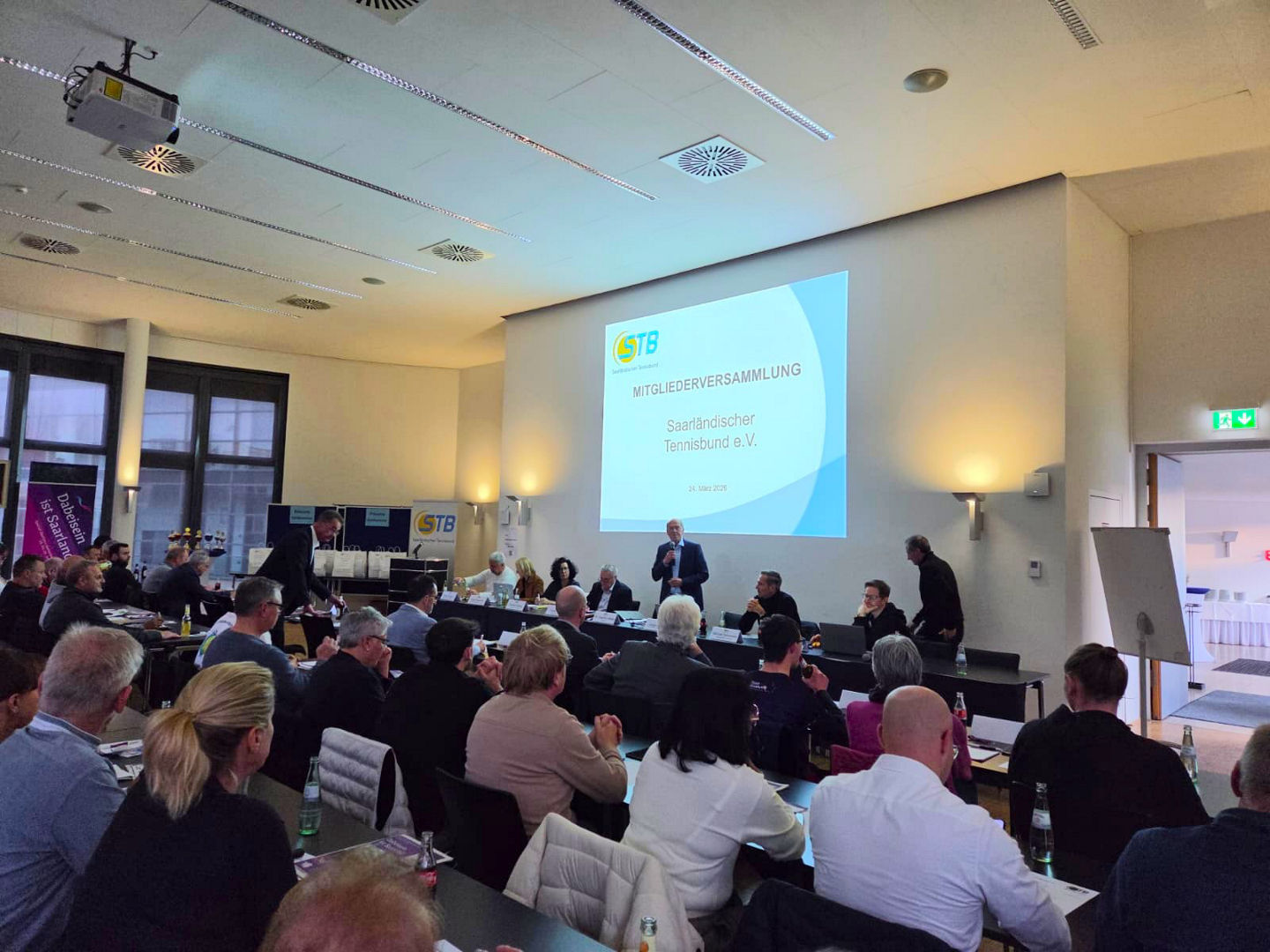 Blick in den Raum der Mitgliederversammlung, auf dem die Teilnehmenden von hinten und das Präsidium am anderen Ende des Raumes vor der Präsentation von vorne zu sehen sind