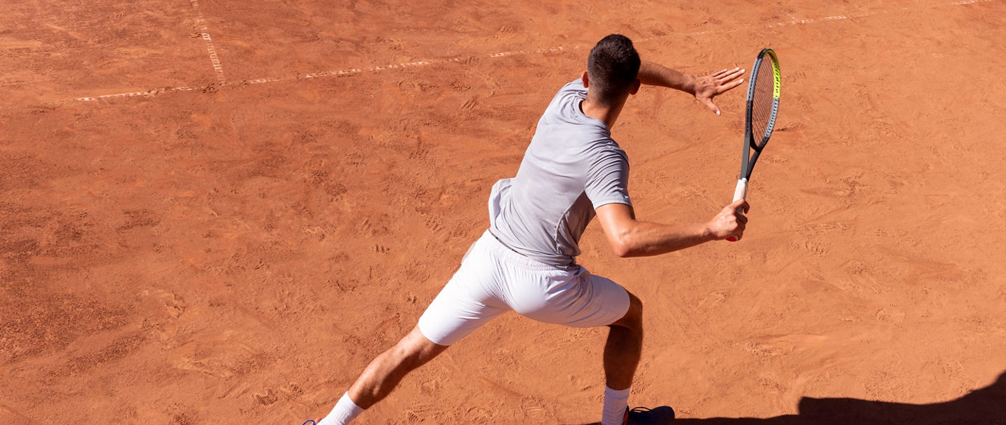 Professional tennis player performs forehand hit on clay tennis court. Young male athlete with tennis racket in action. Junior tennis sport. Back view, shadow, copy space