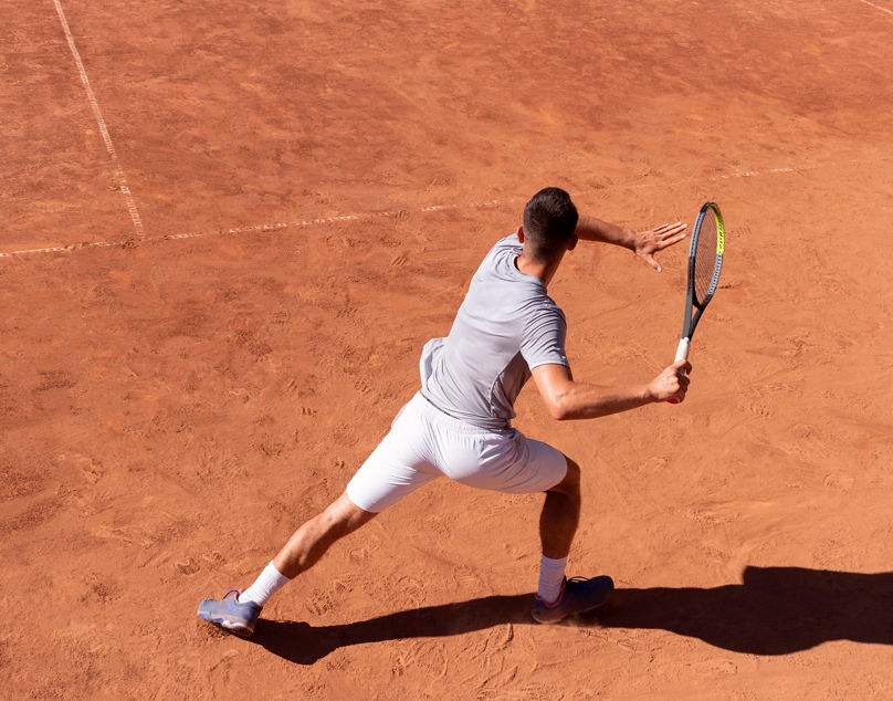 Professional tennis player performs forehand hit on clay tennis court. Young male athlete with tennis racket in action. Junior tennis sport. Back view, shadow, copy space