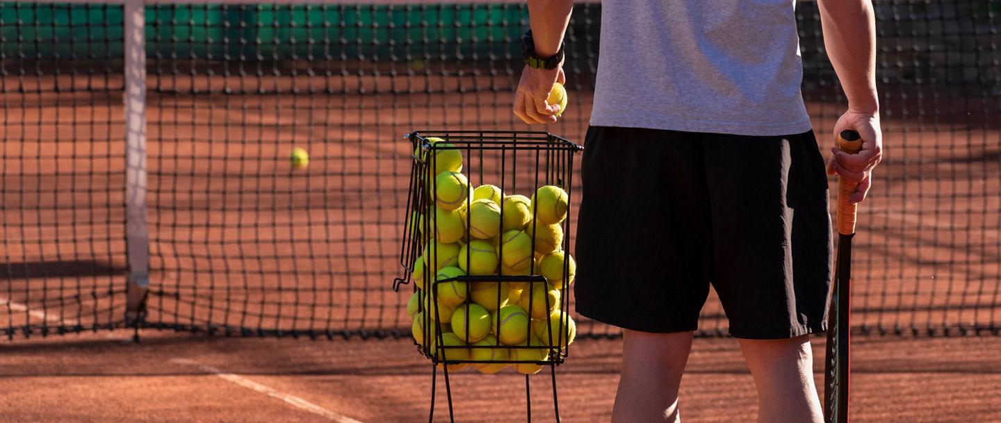 Tennis coach conducts training on red clay court, basket with tennis balls near him. Blurred background. Sports activity and leisure concept. Lifestyle