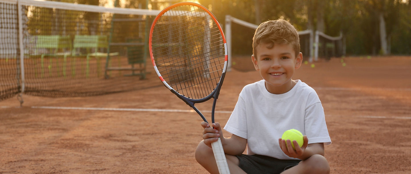 Kleiner Junge sitzt mit Schläger und Ball in der Hand auf einem Tennisplatz