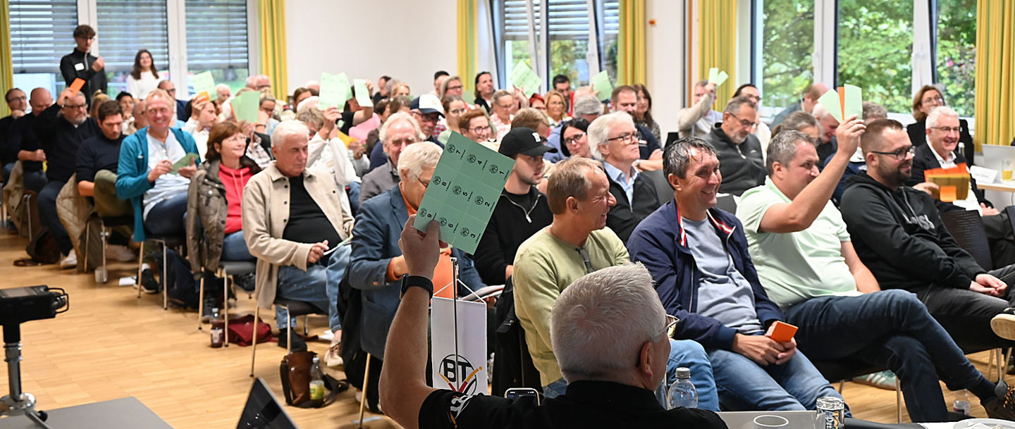 Voller Veranstaltungsraum mit sitzenden Menschen, die teilweise Stimmkarten in die Höhe halten