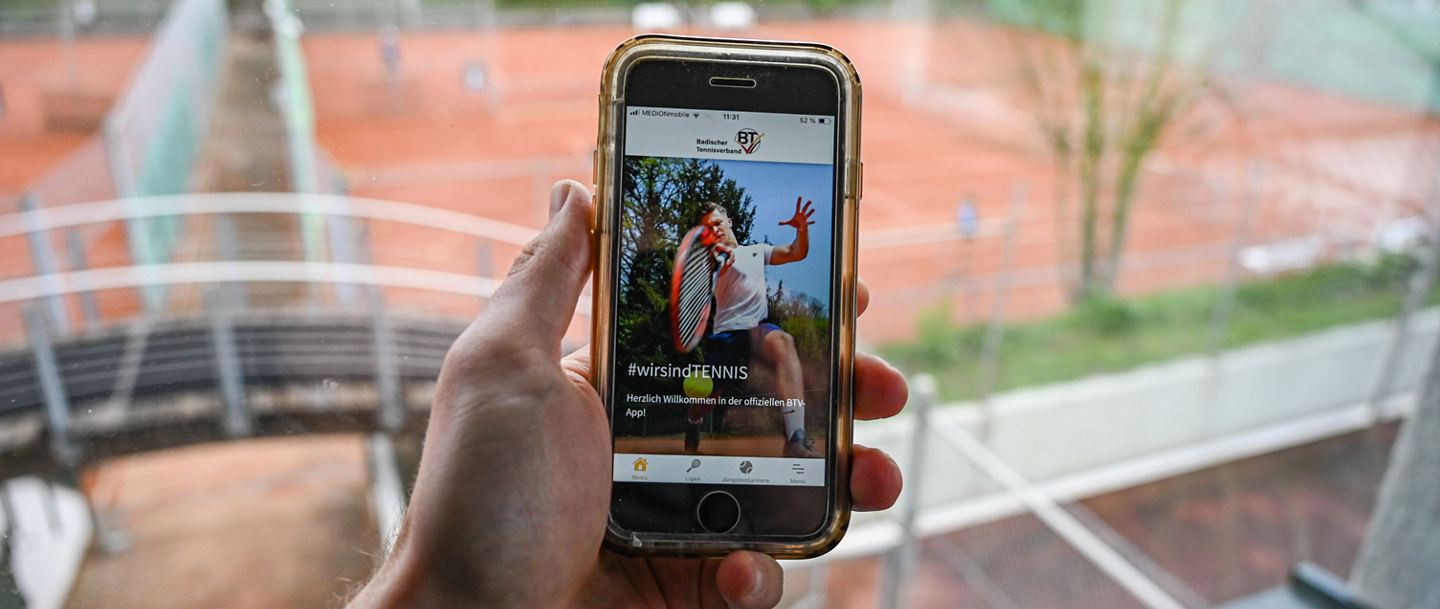 Hand mit Handy in der Hand und Tennisplätzen im Hintergrund