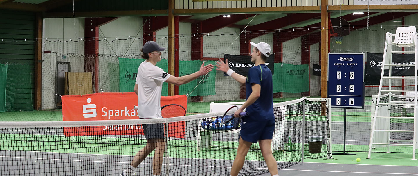 Zwei Spieler kommen auf einem Platz in der Tennishalle am Netz zusammen und klatschen sich ab
