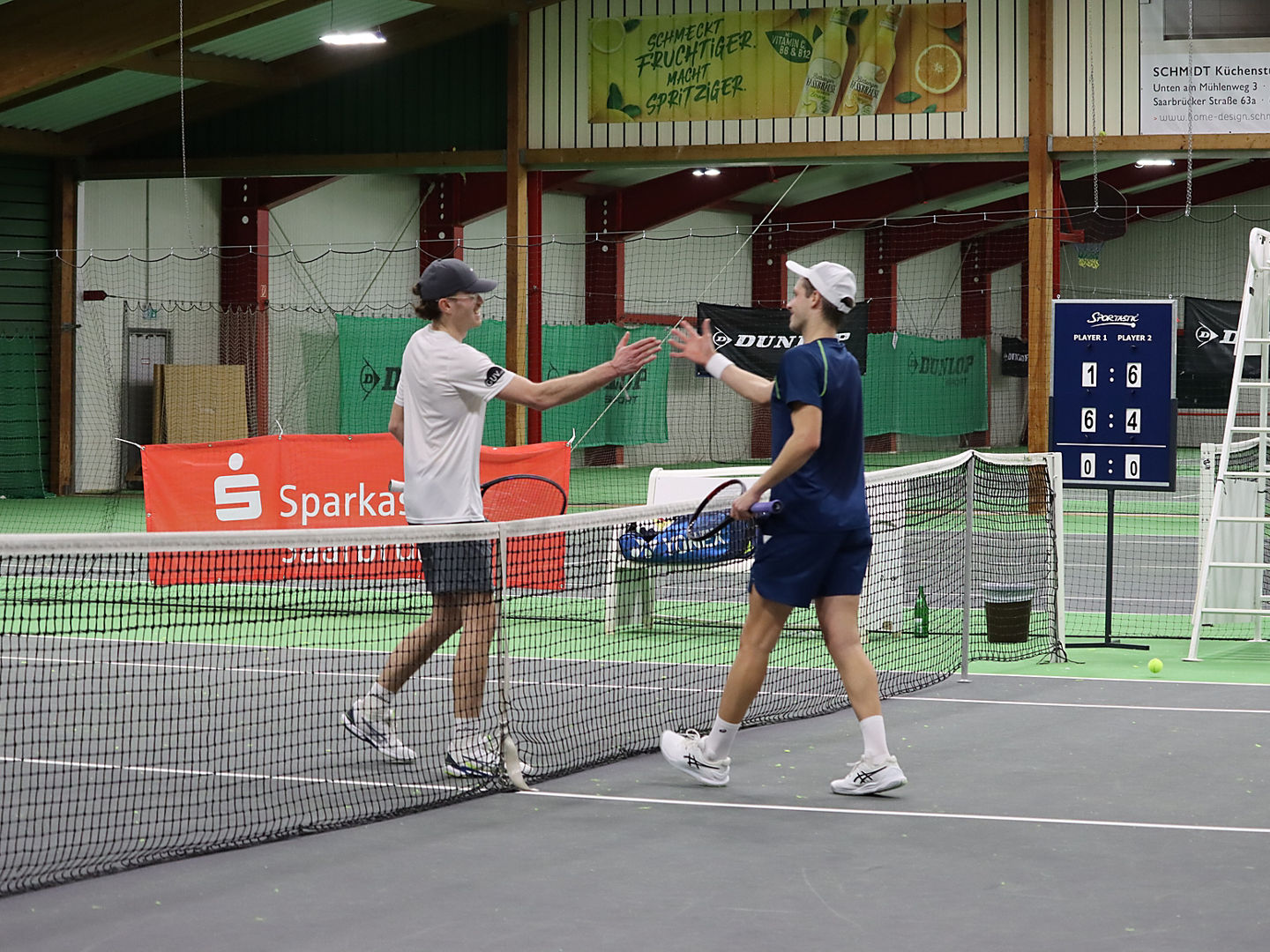 Zwei Spieler kommen auf einem Platz in der Tennishalle am Netz zusammen und klatschen sich ab