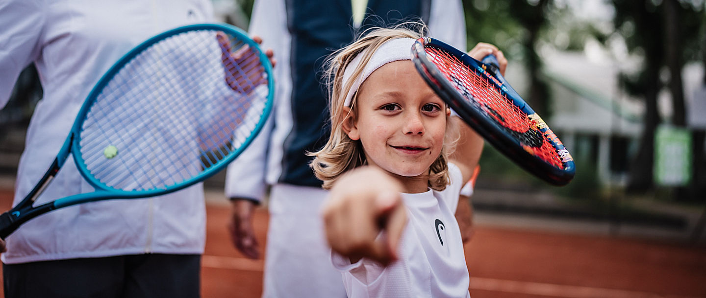 Kind zeigt in die Kamera und hält Tennisschläger in der Hand