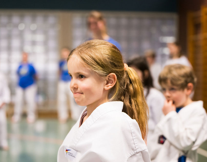 Mädchen in weißem Karateanzug steht in Sporthalle, weitere Kinder in Kampfsportkleidung im Hintergrund.