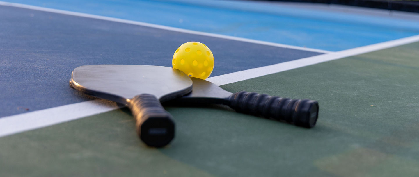Pickleball paddles and a yellow ball on a court with a net in the background, captured during sunset.